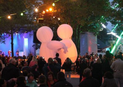 Aufblasbare mit Helium gefüllte Ballonfiguren, bunte Lichterketen hängen in den Bäumen, auf der Bühne spielt eine Band, Zuschauer sitzen auf Bänken im Publikum