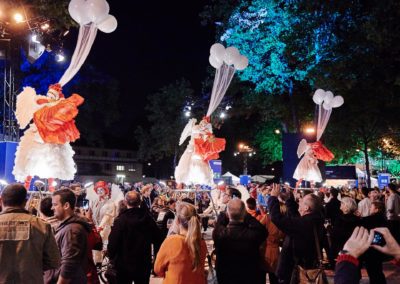 Artisten mit pompösen Kleidern haben mit Helium gefüllte Luftballons an ihren Hüten und stehen auf Podesten und tanzen, Zuschauer sehen zu