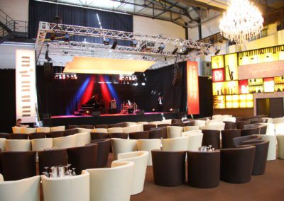 Bühne mit Klavierspieler, Kronleuchter hängen von der Decke, Loungesessel mit Tisch für Zuschauer, links und rechts beleuchtete Bühnenkulisse Bar