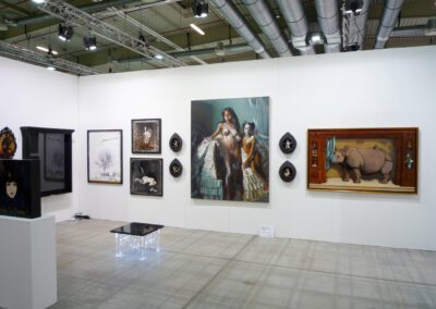 Blick auf eine Kunstwand der Art Fair Messe in Köln, an der Wand hängen dunkel gehaltene Bilder und andere Kunstwerke, in der Mitte vom Raum steht ein kleiner beleuchteter Tisch