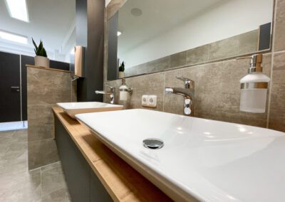 WC, weiße Porzellan Waschbecken, Unterschrank Waschbecken mit schwarzen Fronten und Eicheholzplatte, Spiegel, Wand- und Bodenfliesen in Betonoptik, WC Kabinen