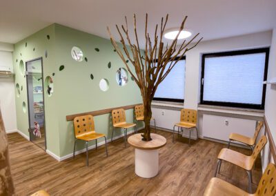 Wartebereich für Kassenpatienten mit einem Baum in der Raummitte und Stühlen aus Holz